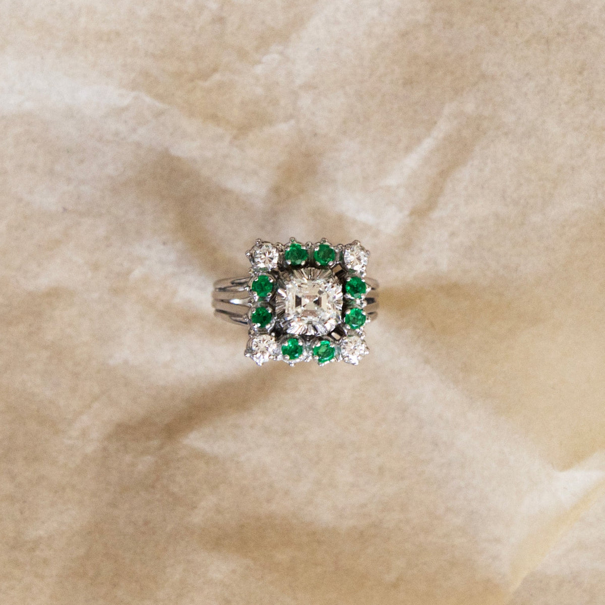 Bague marguerite en or gris 18 carats sertie d'un diamant taille émeraude entouré d'émeraudes et de diamants, vue de face sur fond beige.