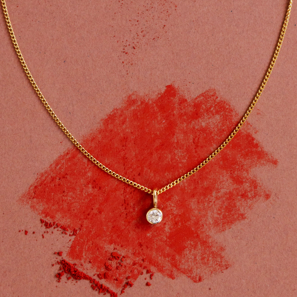 Pendentif diamant de 0.1 carat sur une chaine en or jaune 18 carats maille gourmette, sur un fond rouge.