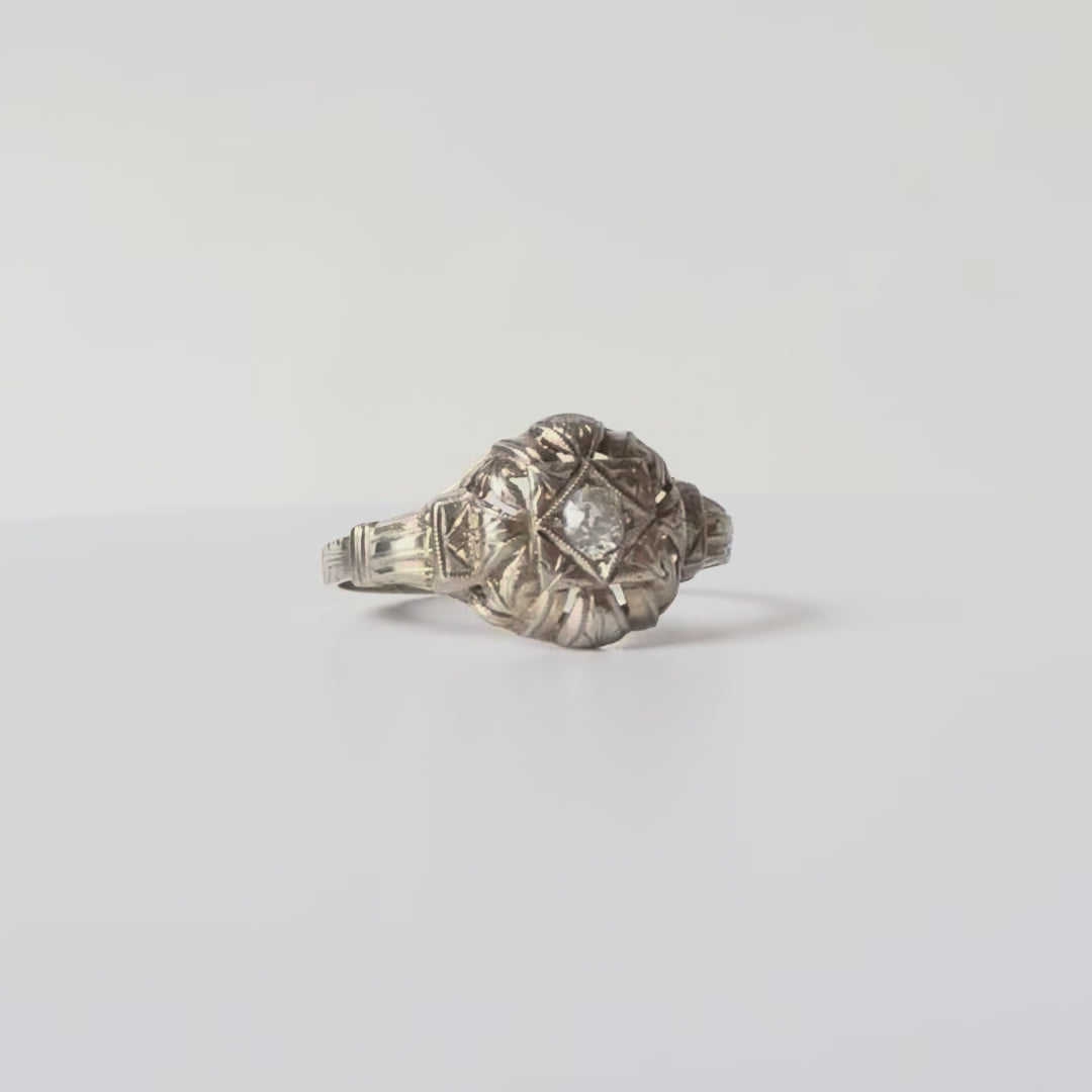 Élégante bague en or blanc 18 carats, sublimée par un diamant serti clos. Sa monture délicatement ajourée est décorée de motifs feuillagés et d’arabesques gravées, offrant un charme raffiné et intemporel.